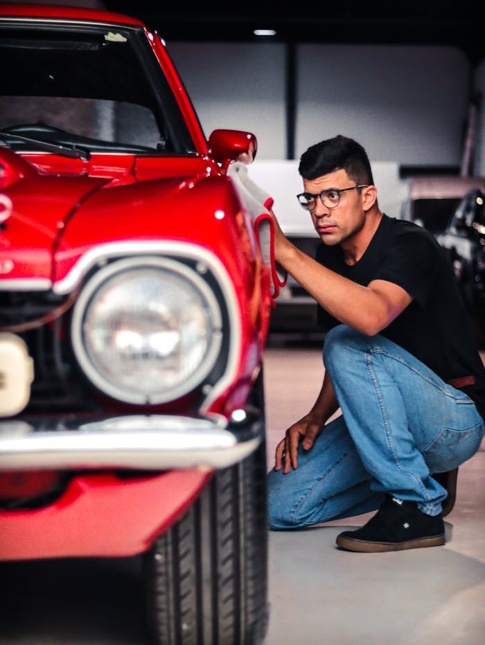 A man kneels to inspect a classic red car in a garage setting, emphasizing detail and care.
