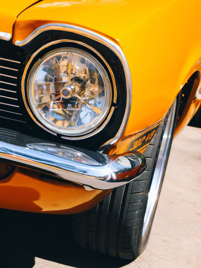 contact-img Close-up of a classic yellow car's headlight with chrome details, shot in bright sunlight.
